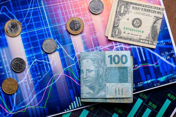 Gráfico ao fundo, enquanto na frente possui dolares e reais, bem como moedas brasileiras e americanas.