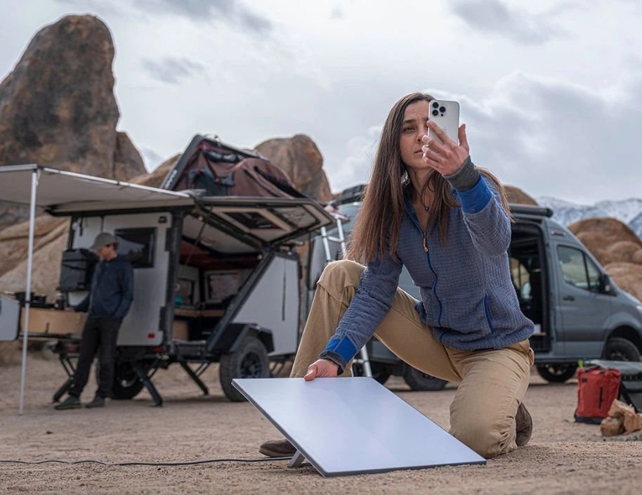 Um casal em um ambiente desértico acampado próximo a uma van. Ela segura um celular em uma das mãos, tentando conexão com a internet, e uma antena da Starlink em outra, instalada no chão.