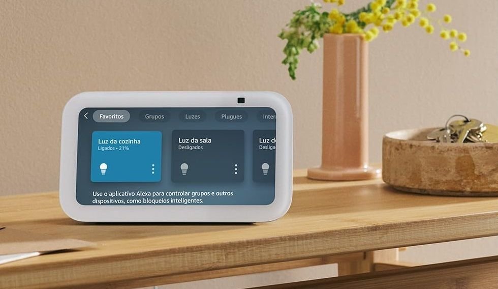 Um Amazon Echo Show de 5ª Geração em cima de uma mesa de madeira, ligado e com uma tela de gerenciamento de lâmpadas inteligentes. Ao lado, há um vaso de flores e um chaveiro também de madeira.