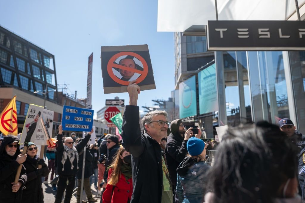 Pessoas protestando contra Elon Musk com cartazes e bandeiras em frente a uma loja da empresa.