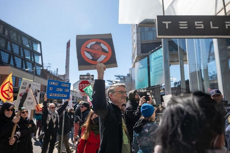 Pessoas em uma passeata contra a Tesla na frente de uma loja da marca. Um homem segura um cartaz com a foto de Elon Musk e um sinal de proibido.