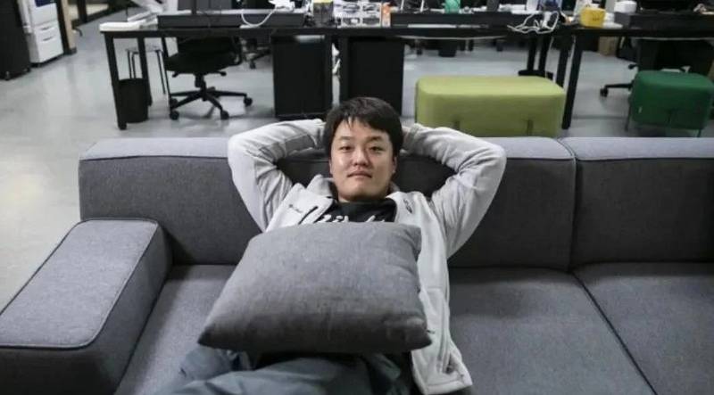 um homem sul-coreano deitado em um sofá cinza, com um escritório ao fundo