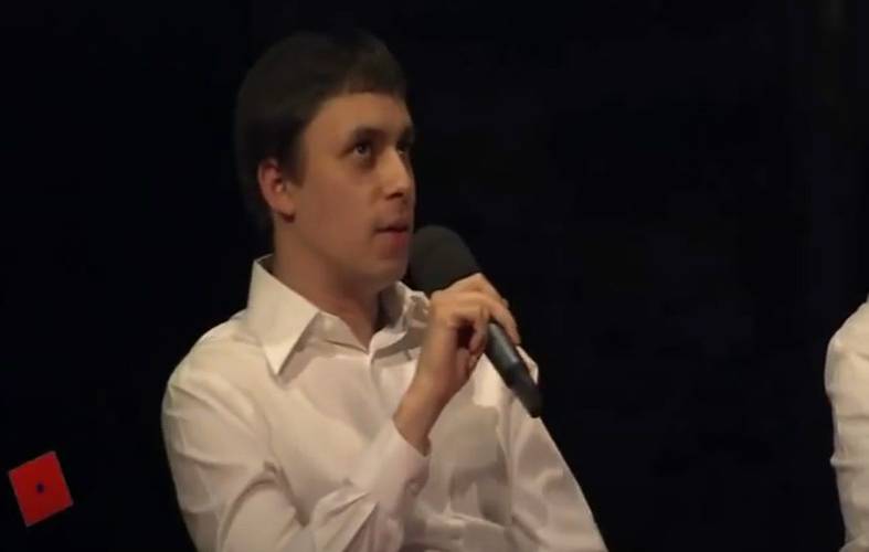 Um homem de camisa branca segurando um microfone e falando em um evento.