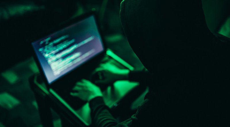 um homem coberto por um capuz e de costas, usando um computador no escuro sob uma iluminação verde