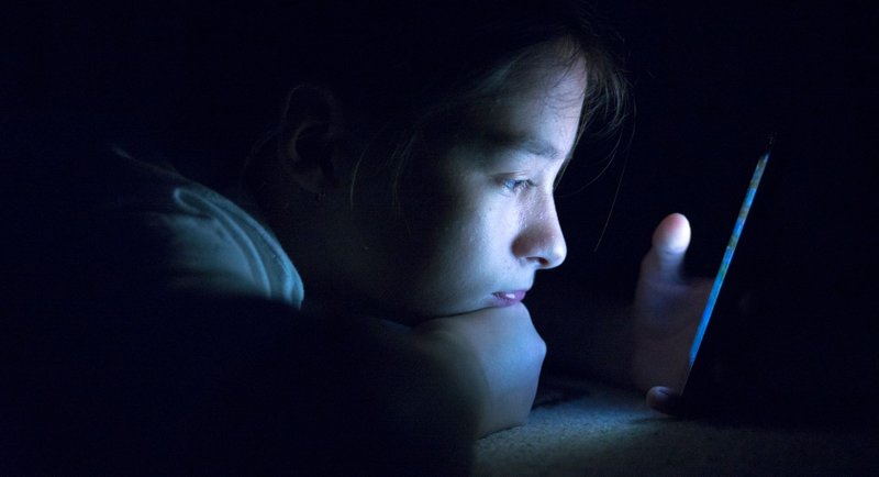 uma criança no escuro deitada de barriga para baixo, com o rosto iluminado pela tela de um celular.