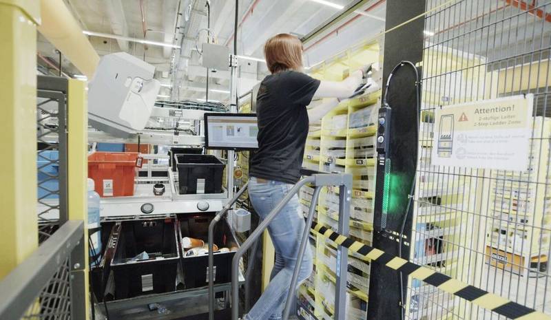 Uma mulher em cima de uma escada tirando objetos de uma pilha de um armazem da amazon