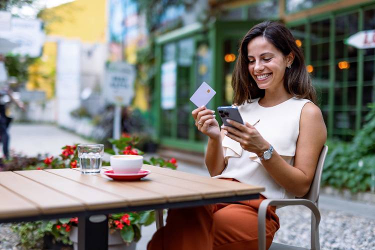 Mulher sentada em frente a uma mesa de cafeteria, com um smartphone e cartão na mão, sorrindo.