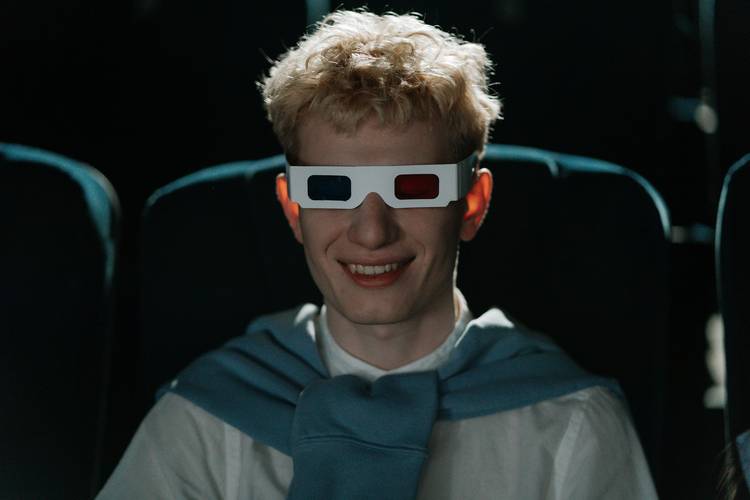 Homem caucasiano usando um áculos 3D de papel e sorrindo dentro de uma sala de cinema escura.